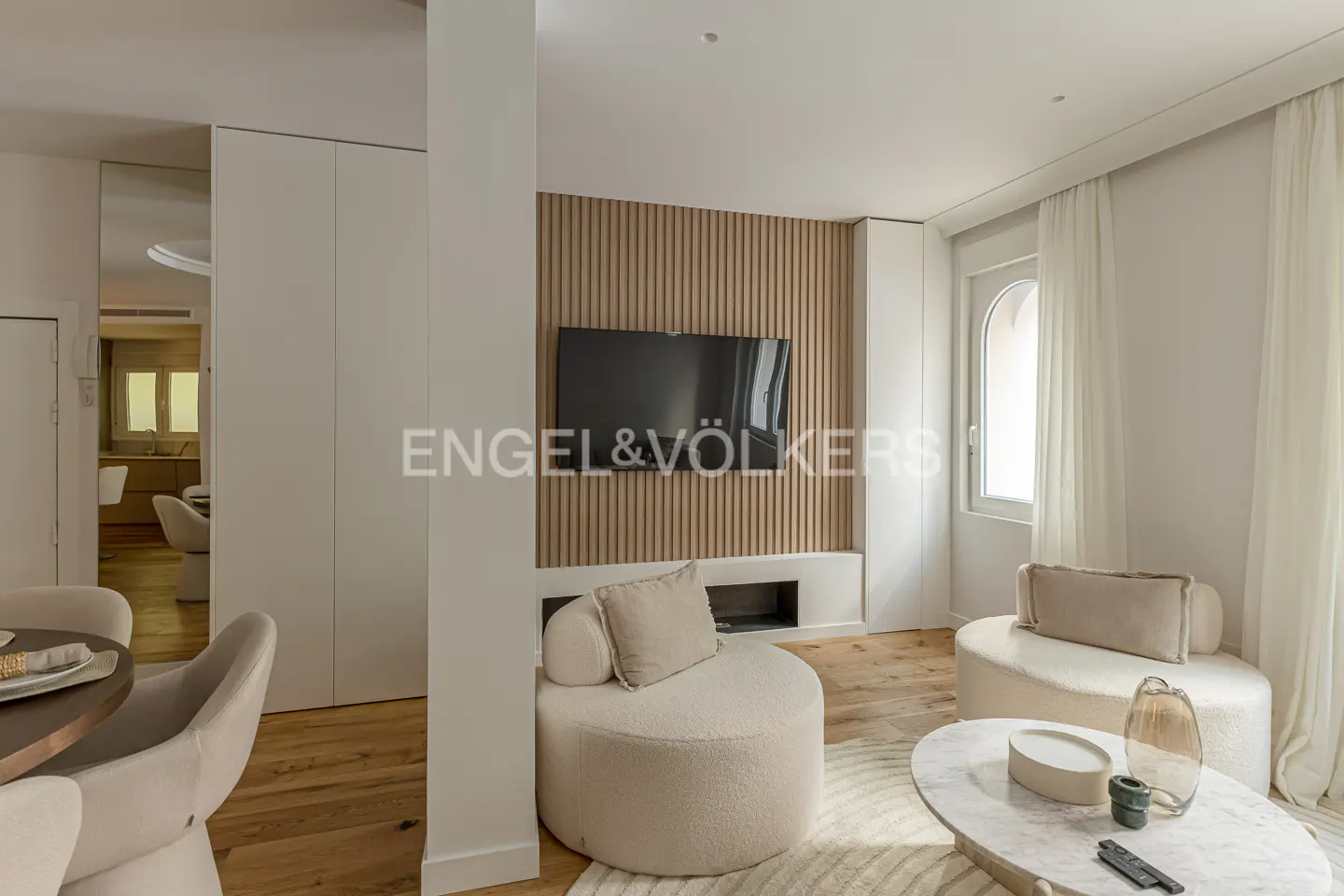 Modern living room with two cream-colored curved chairs, a marble coffee table, and a wall-mounted TV on wood slats.