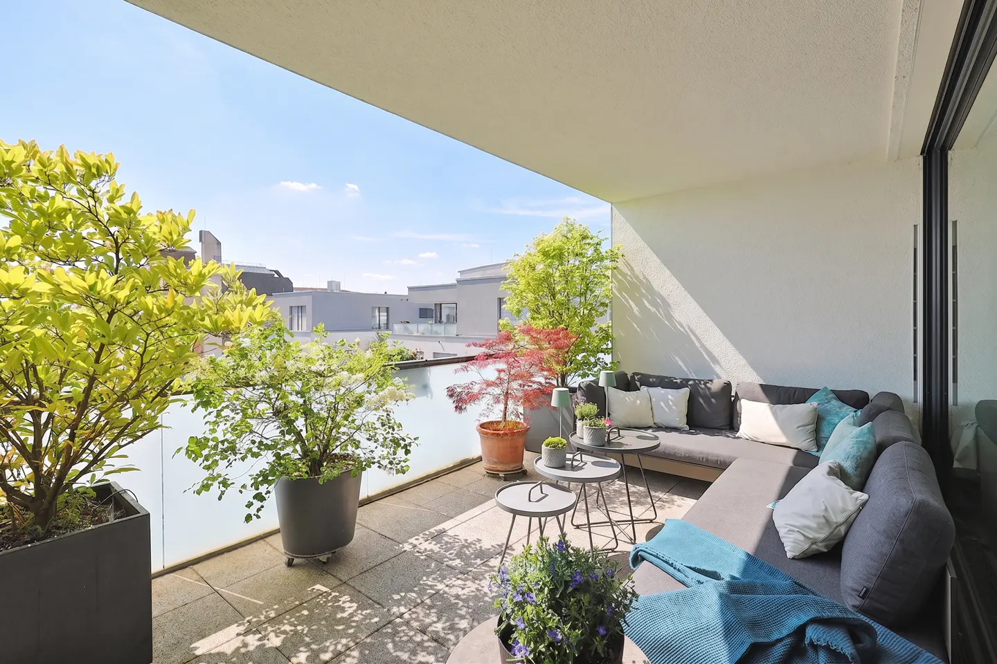 Modern balcony with a gray sectional sofa, potted plants, small tables, and a view of urban buildings under a blue sky.