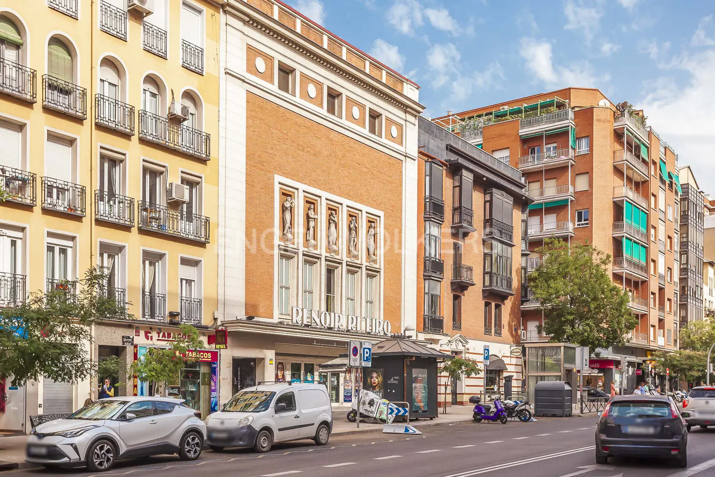 Street view of Madrid, Spain, with cars, buildings, and the Renoir Retiro cinema. The buildings are yellow and brick, with balconies and windows.