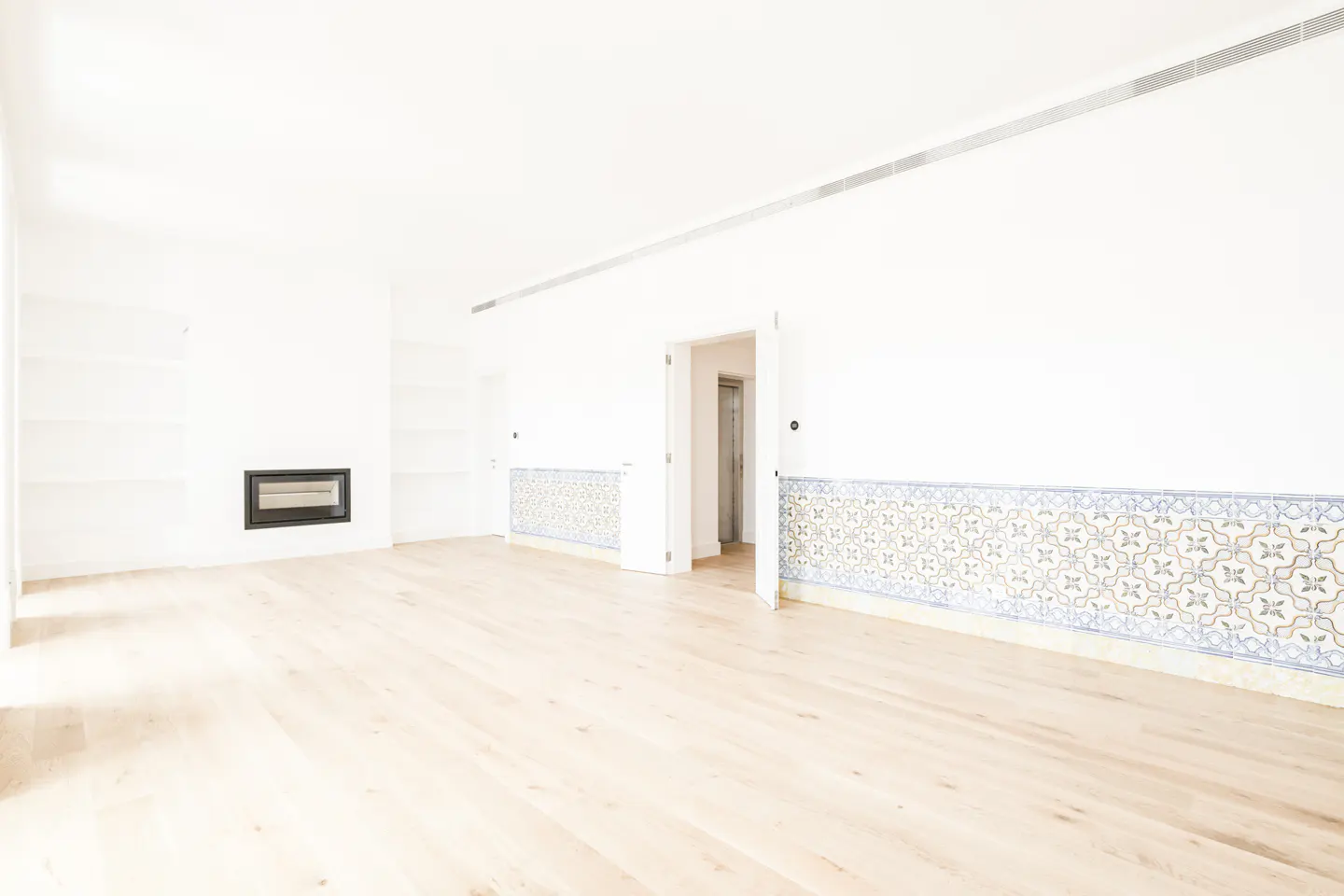 Bright, empty room with light wood floors, white walls, and a black fireplace. Decorative blue and white tiles line the lower walls. An open doorway leads to another room.