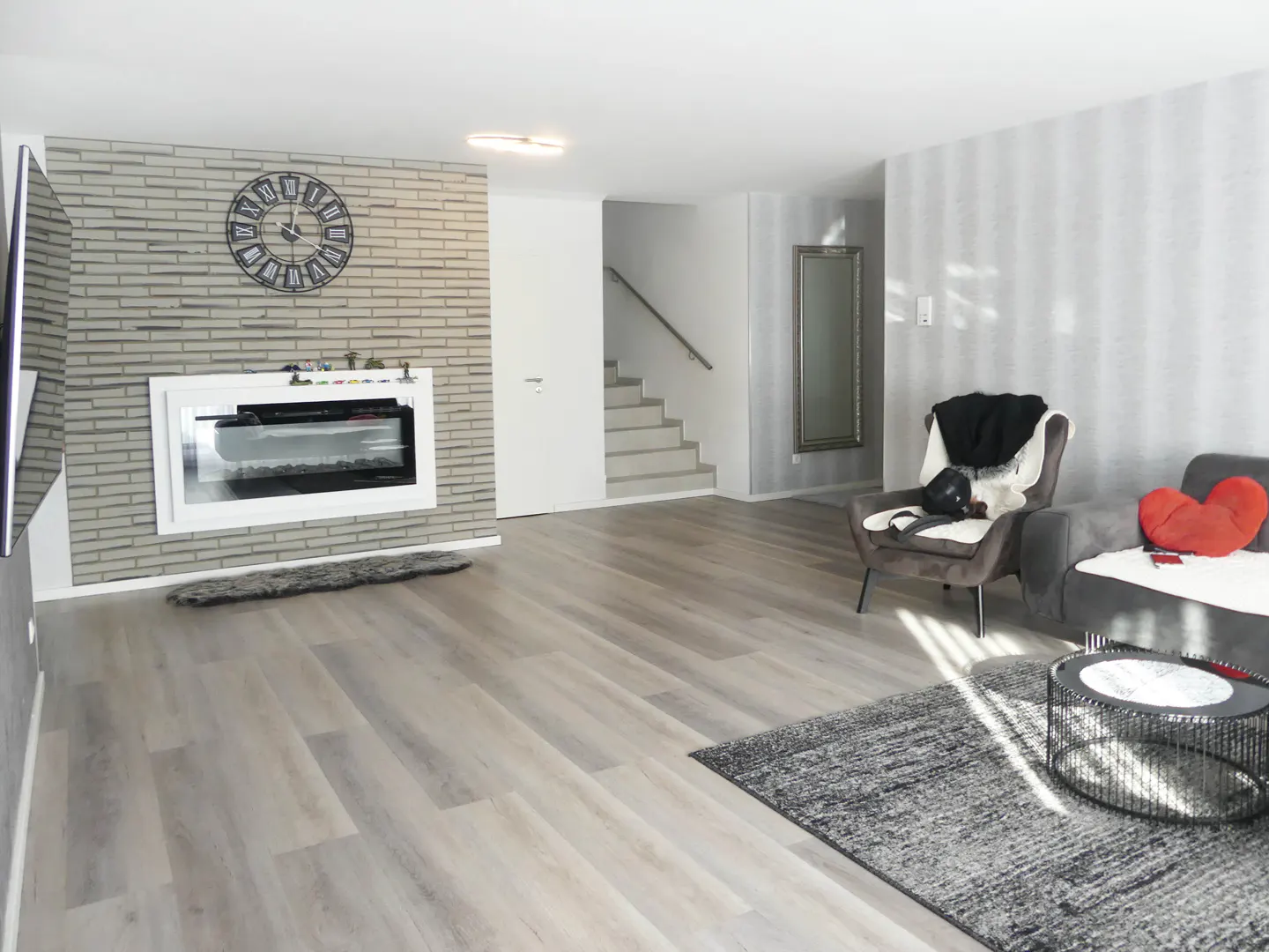 Living room with gray wood floors, brick wall with clock and fireplace, gray sofa and chair, and stairs in the background.