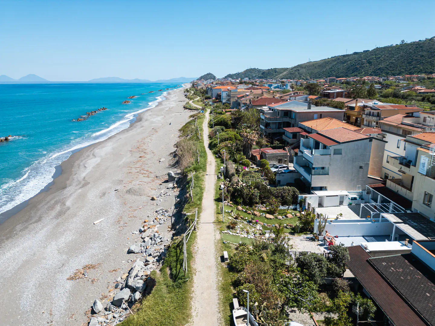 Aerial view of a sandy beach and turquoise sea, with a path leading to houses with red tile roofs. Hills are visible in the background.