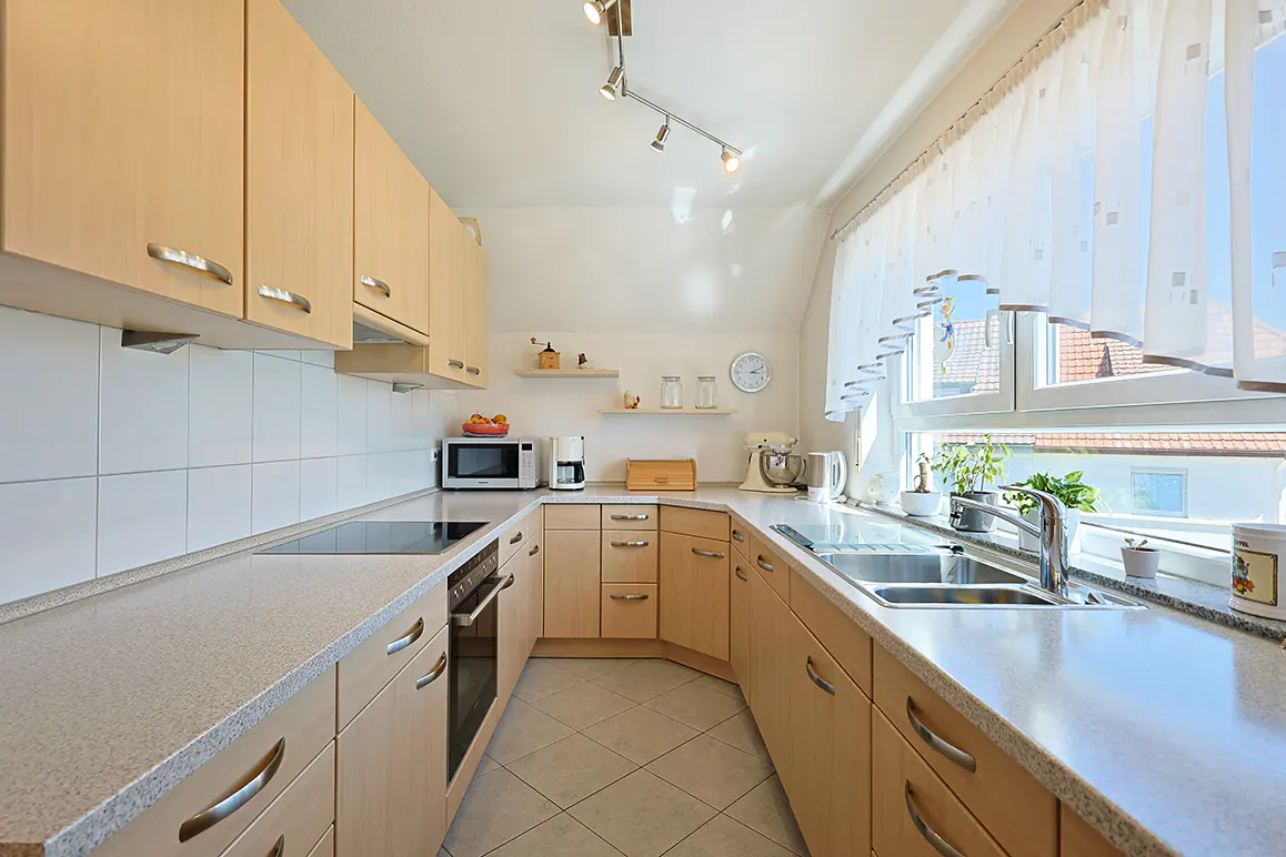 U-shaped kitchen with light wood cabinets, speckled countertops, stainless steel appliances, and a window with white curtains.