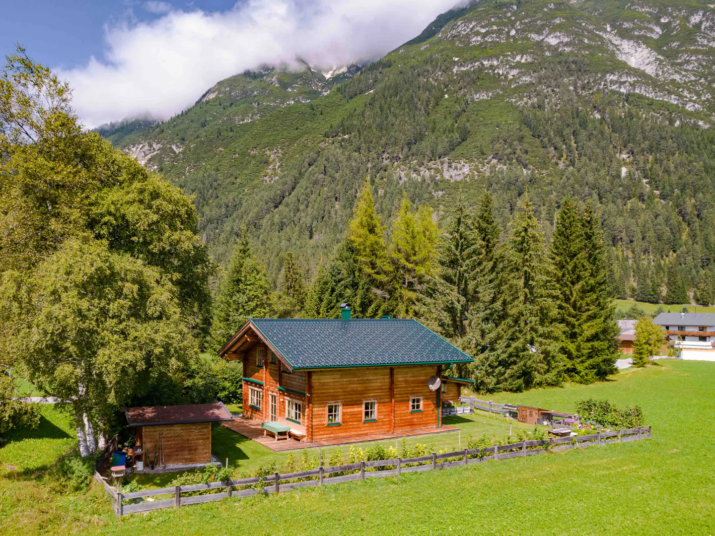 A wooden chalet with a green roof sits in a lush green meadow, backed by a forest and mountains.