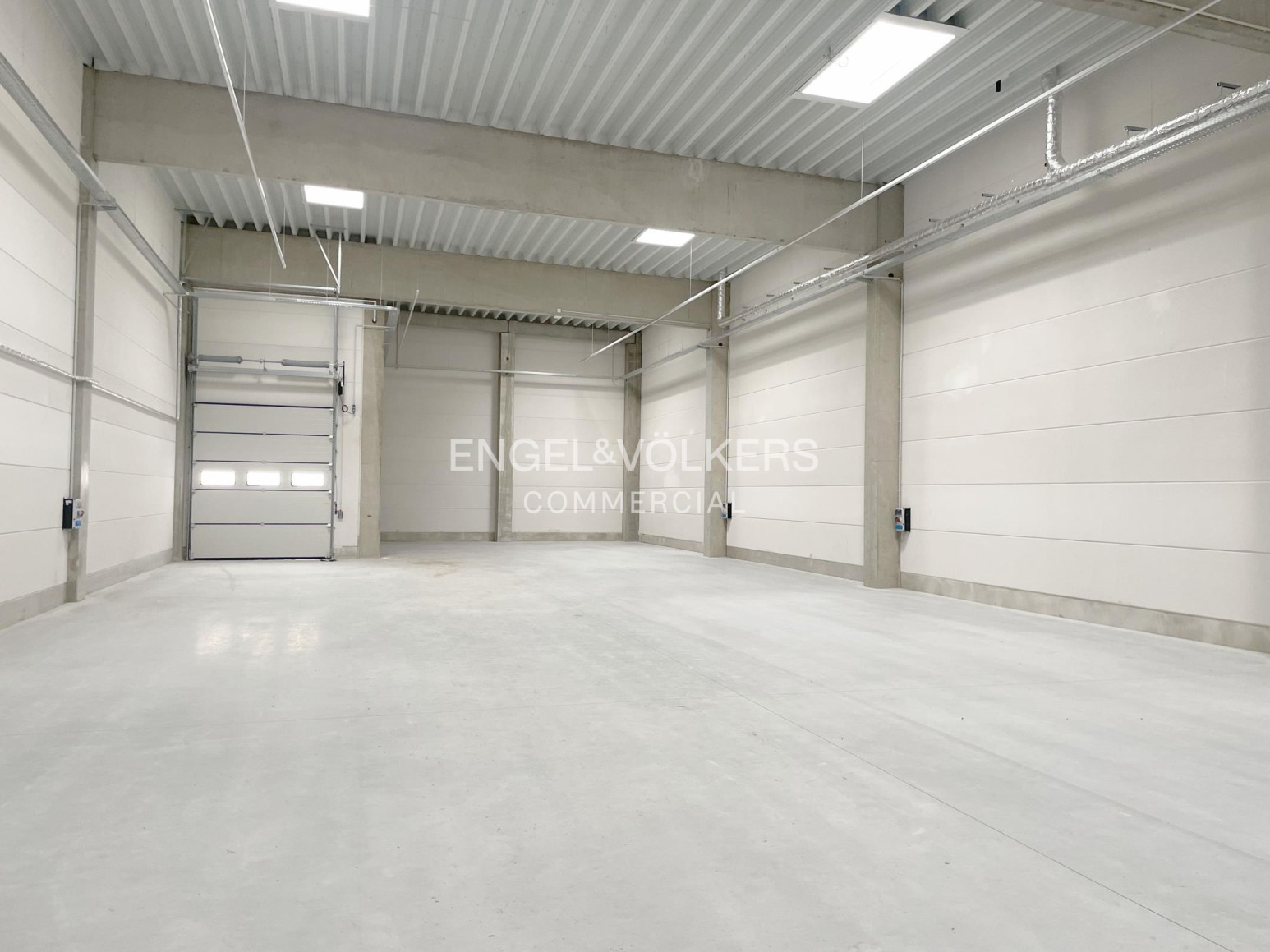 Empty, spacious industrial warehouse with white walls, concrete floors, high ceilings, and a large roll-up garage door. [AI-generated text]