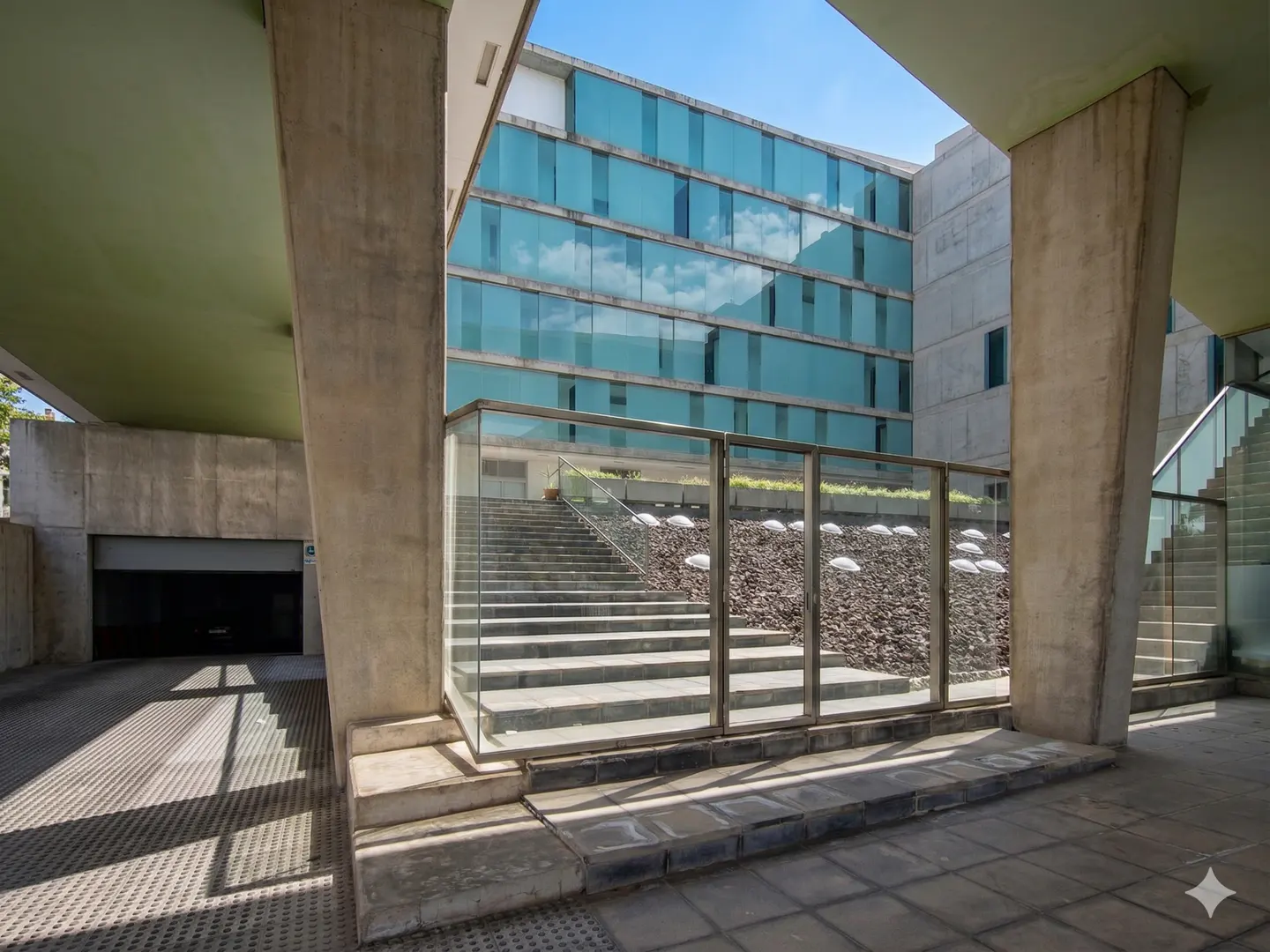 Modern building exterior with concrete pillars, glass facade, and stone stairs leading to a landscaped terrace.