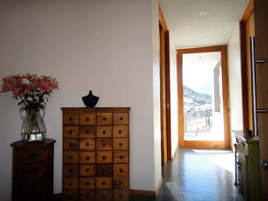 Hallway with a wooden cabinet, black vase, and pink lilies. A doorway leads to a bright view of a town and mountains.