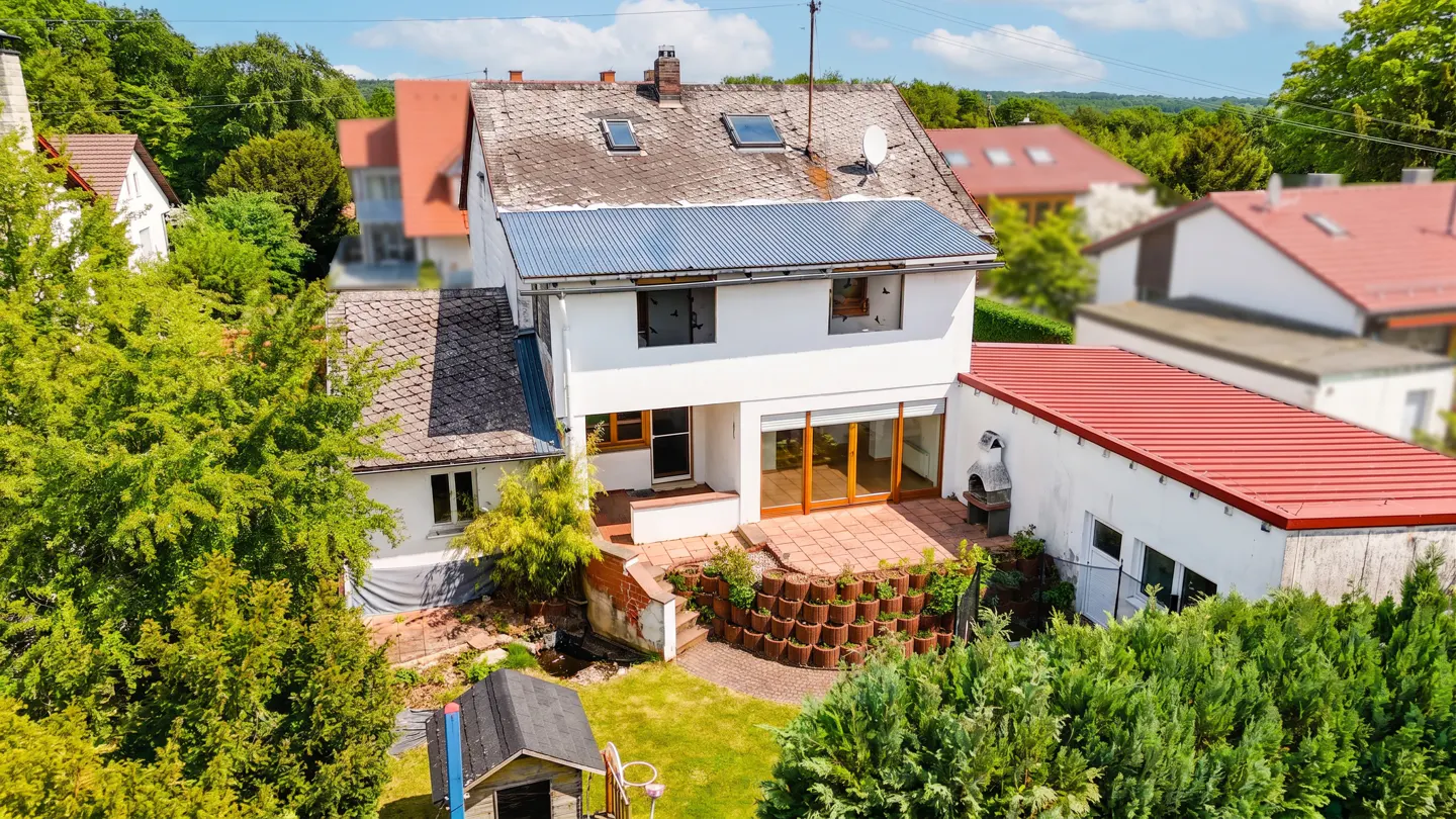 Aerial view of a white two-story house with a gray roof, a brick patio, and a green lawn surrounded by trees.