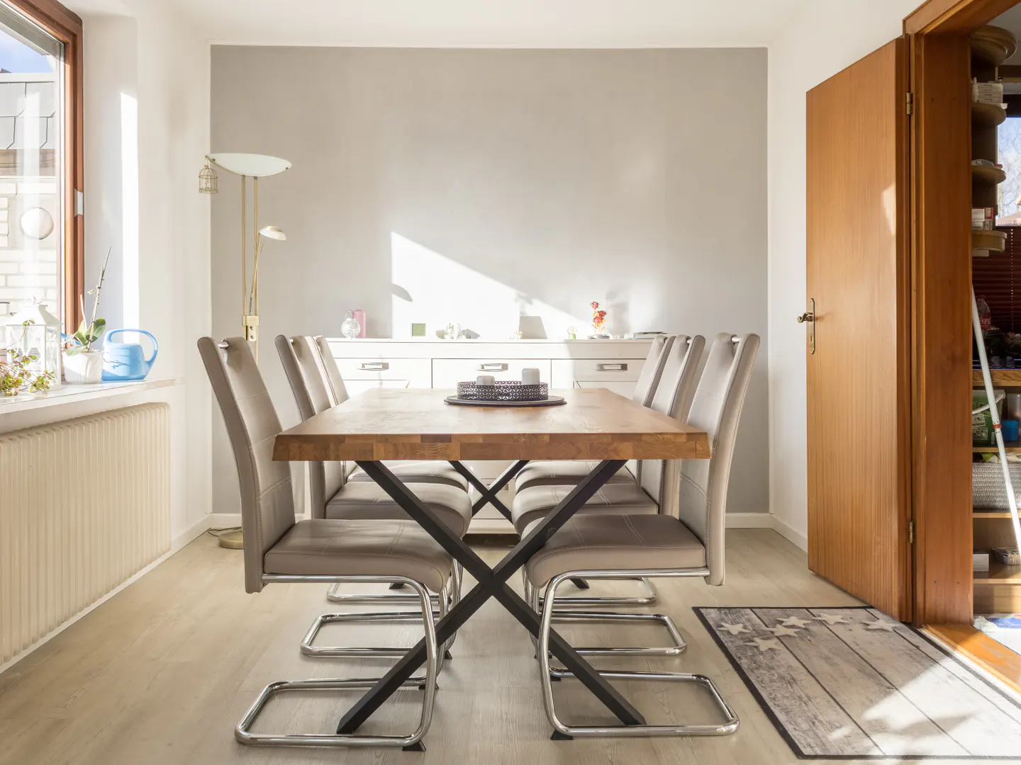 Bright dining room with a wooden table, six gray chairs, and a white sideboard against a gray wall. A wooden door is slightly open.
