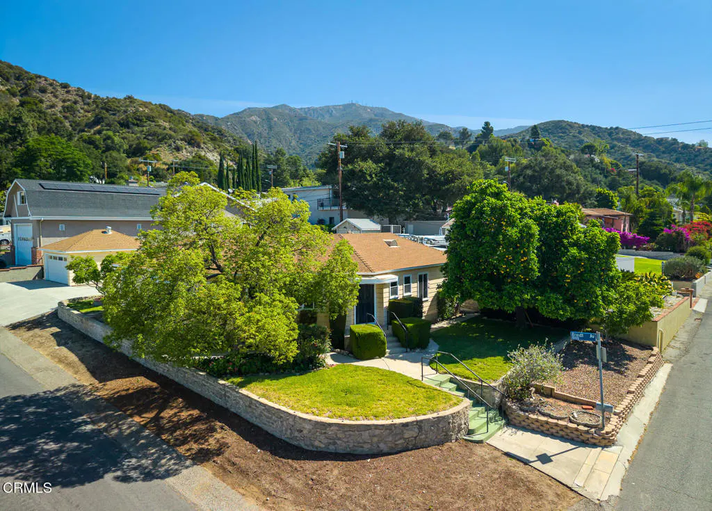 A tan house with a brown roof sits on a green lawn, surrounded by trees and mountains under a clear blue sky.
