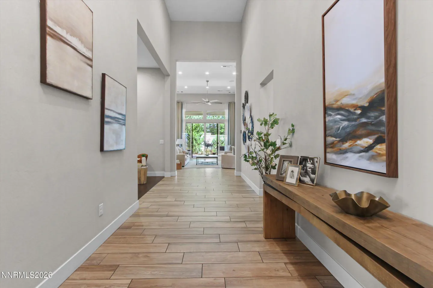 Hallway with light gray walls, wood-look tile floor, and a long wooden bench with art, photos, and a gold bowl. Living room visible in the background.