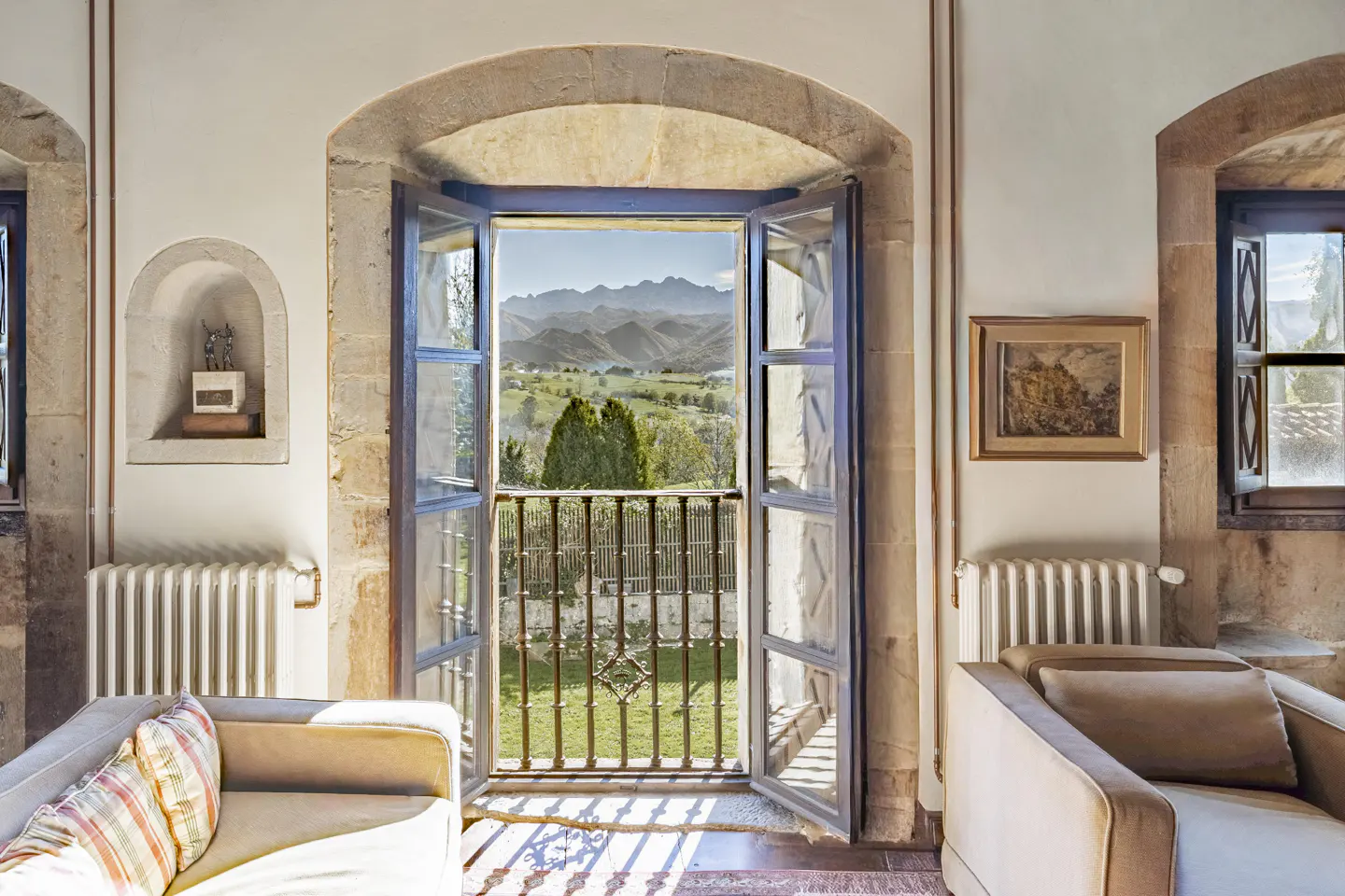 Open French doors in a stone-walled room reveal a balcony overlooking a lush green valley and distant mountain range.