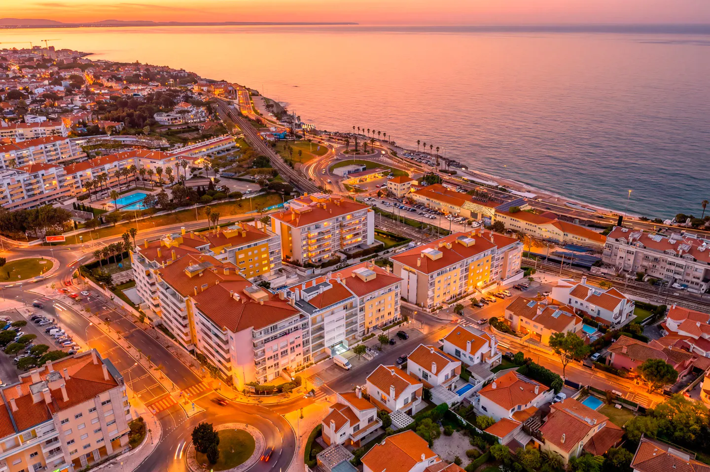 Aerial view of a coastal city at sunset, featuring buildings with red tile roofs and the ocean in the background.
