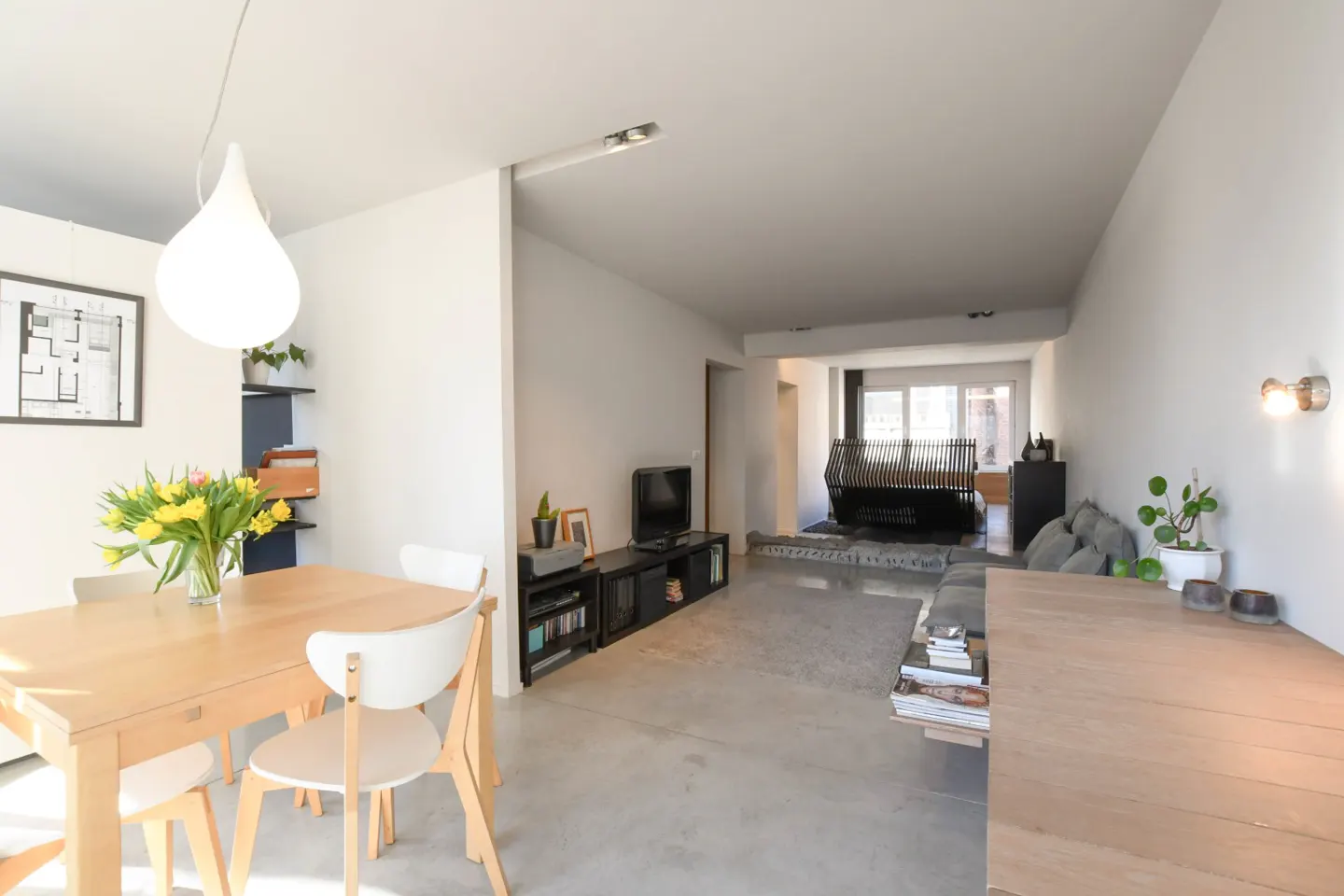 Bright, modern living space with a dining table, chairs, and a vase of yellow tulips. A TV and sofa are visible in the background.