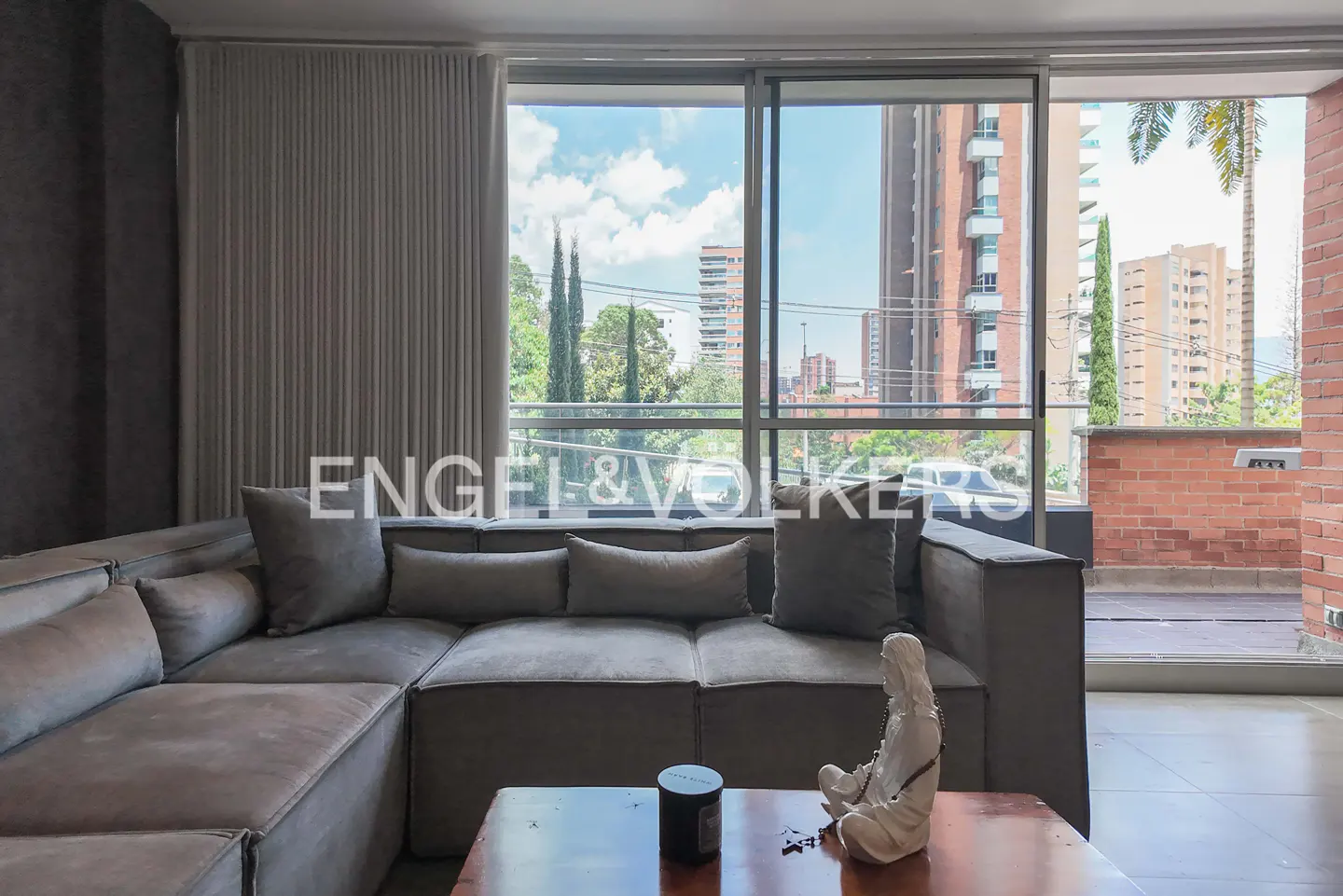 Modern living room with a large grey sectional sofa, a wooden coffee table, and a balcony view of city buildings and trees.