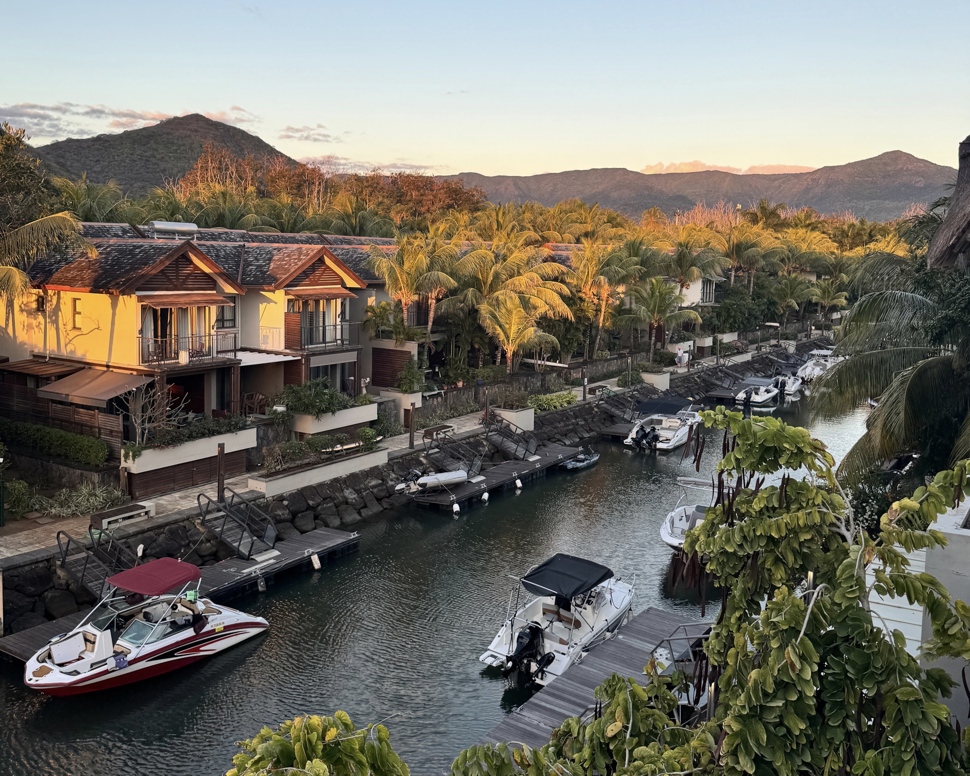 Luxury waterfront villas with private boat docks and moored motorboats at sunset, surrounded by palm trees and mountains. [AI-generated text]