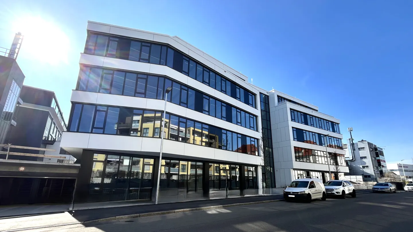 Modern white office building with large windows reflecting the sky, parked cars, and a sunny blue sky.