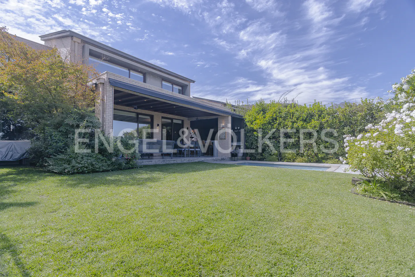 Backyard view of a two-story house with a pool, green lawn, and Engel & Volkers logo.