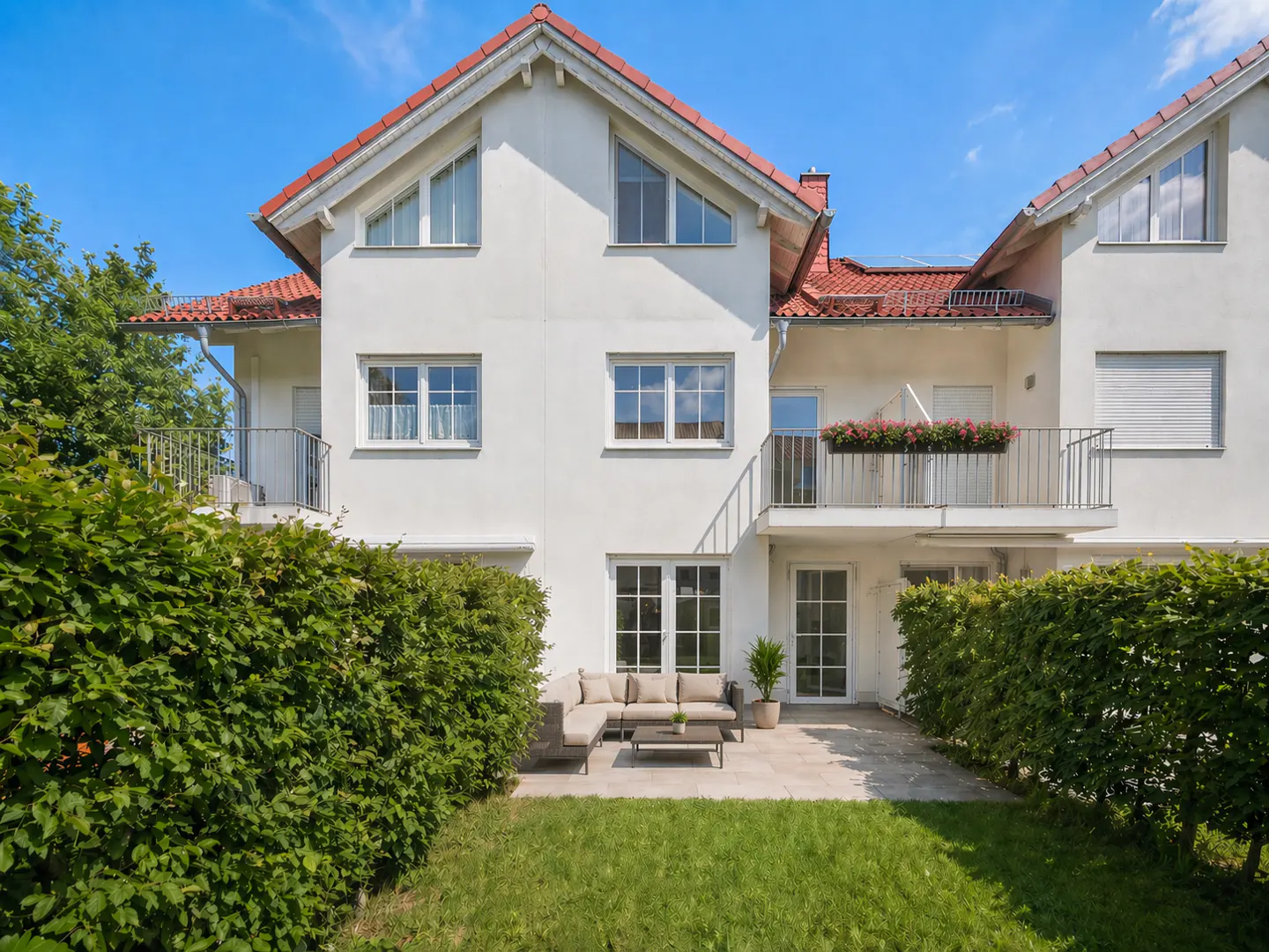 Exterior view of a white two-story house with a red roof, a patio with outdoor furniture, and a green lawn.