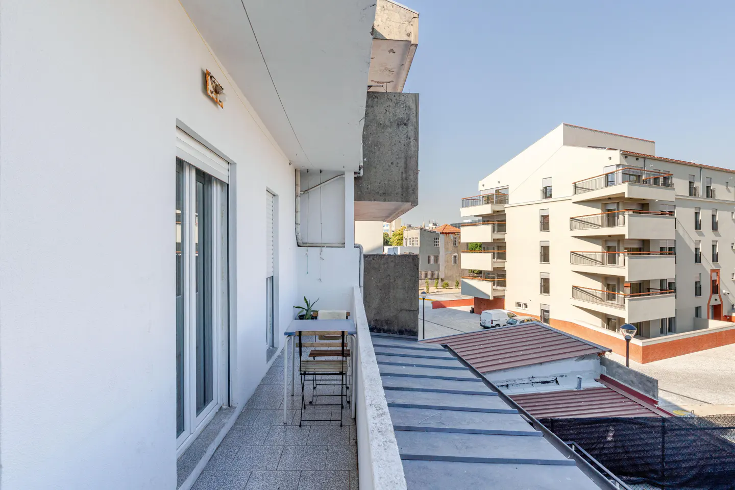 Balcony view with a small table and chairs, white walls, and a view of a nearby building under a clear blue sky.