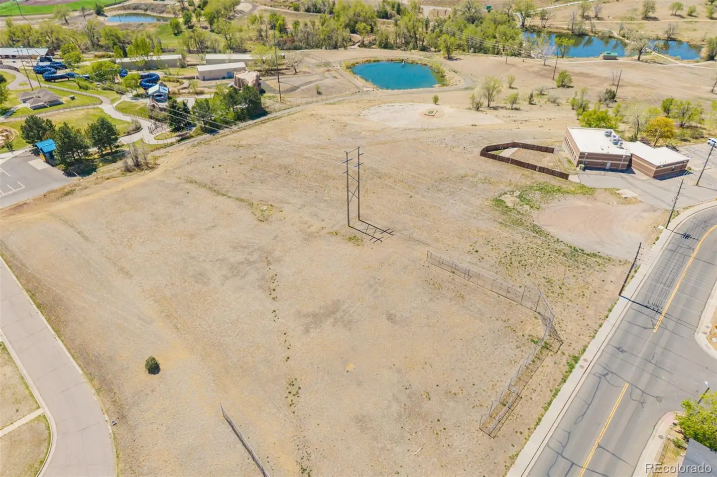 Aerial view of a large, vacant, sandy lot with power lines, a fence, and a building in the background.