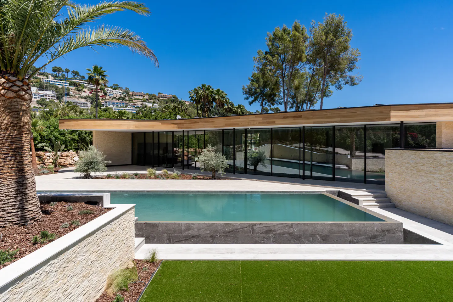 Modern home exterior with a rectangular pool, palm tree, and floor-to-ceiling windows under a clear blue sky.