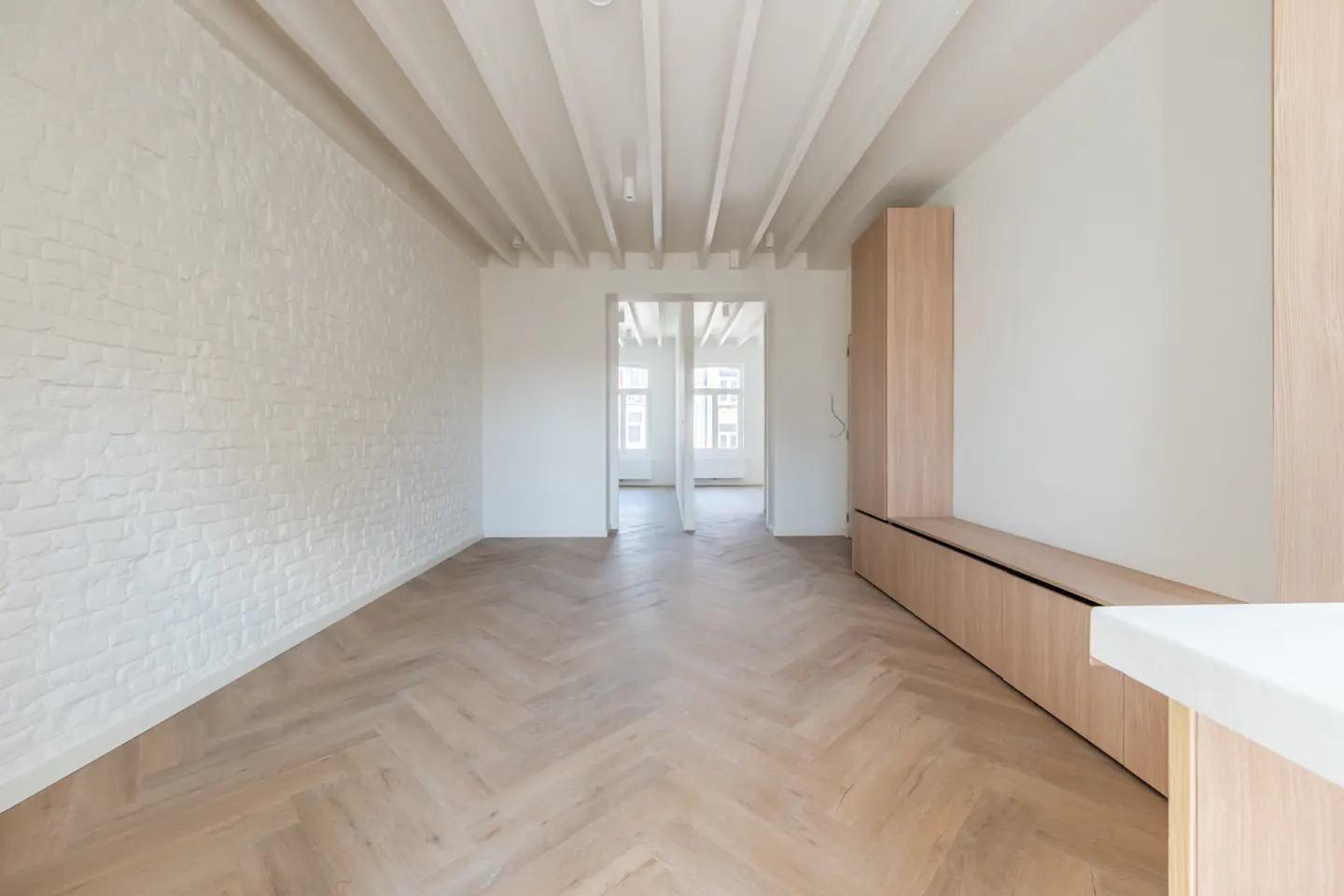 Bright, empty room with herringbone wood floors, white brick wall, and exposed ceiling beams. A doorway leads to another room with windows.