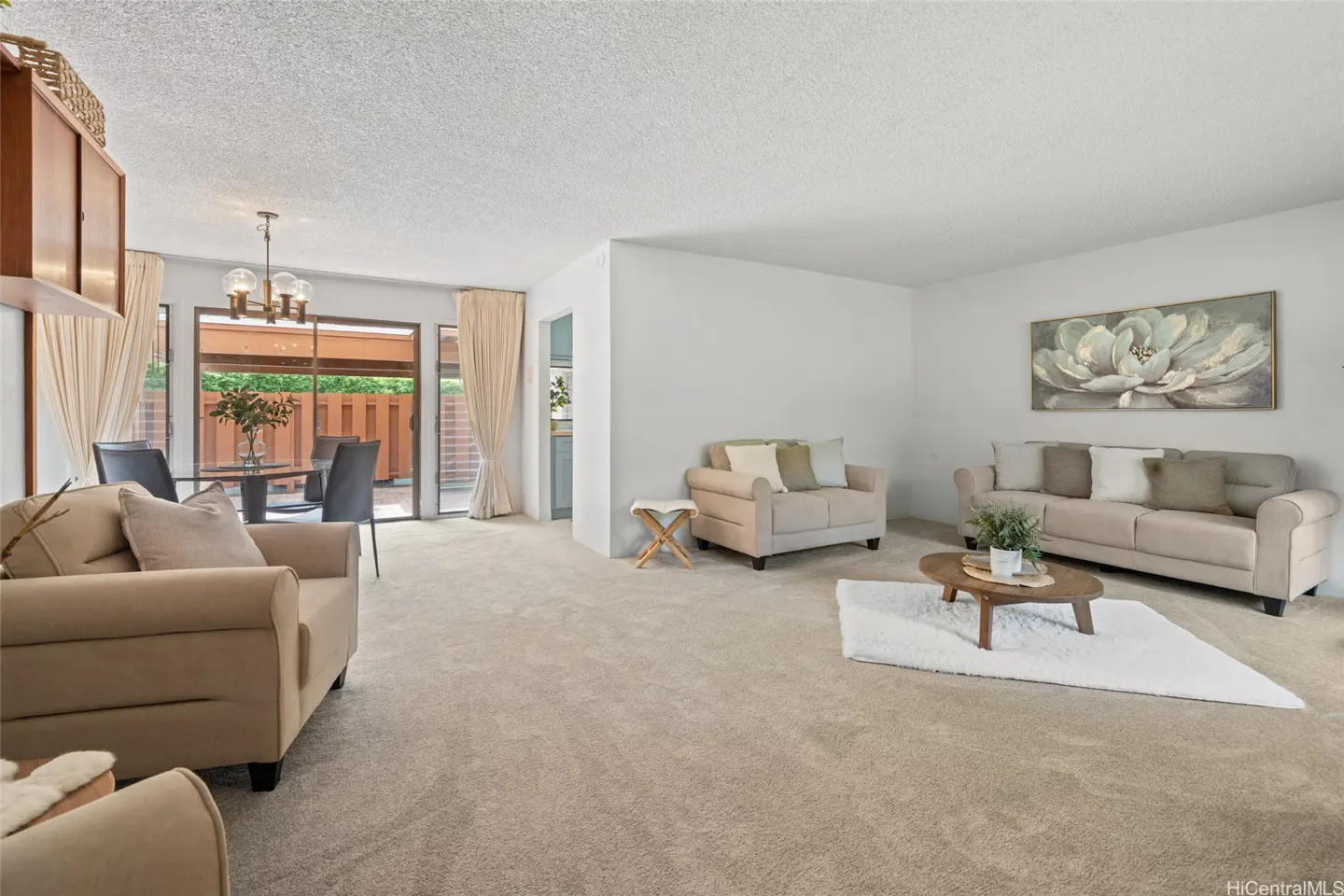 A bright living room with beige carpet, white walls, and neutral furniture. A dining area is visible through sliding glass doors.