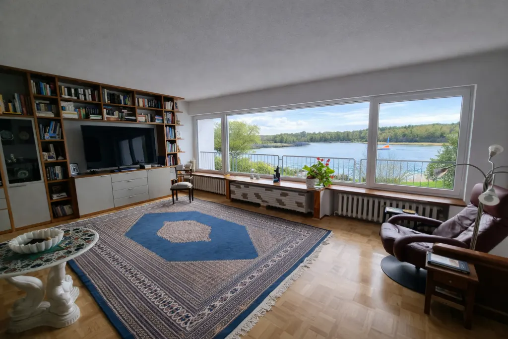 A living room with a large window overlooking a lake. A blue rug covers the wooden floor. A bookcase and TV are on the left.