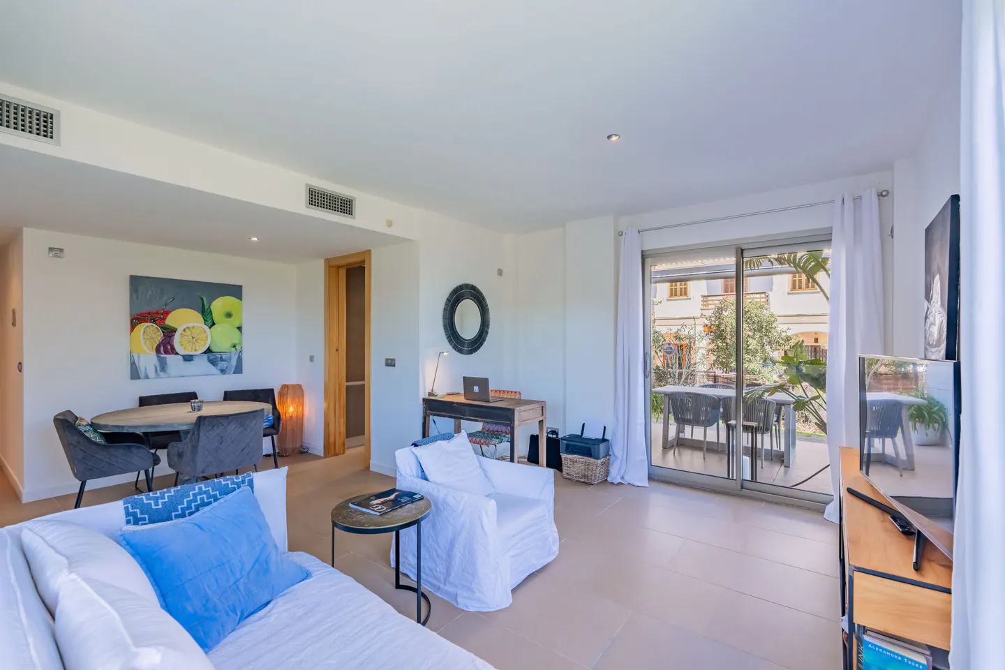 Bright living room with white sofa, blue pillows, dining table, desk, and sliding glass doors to a patio. Fruit painting on the wall.