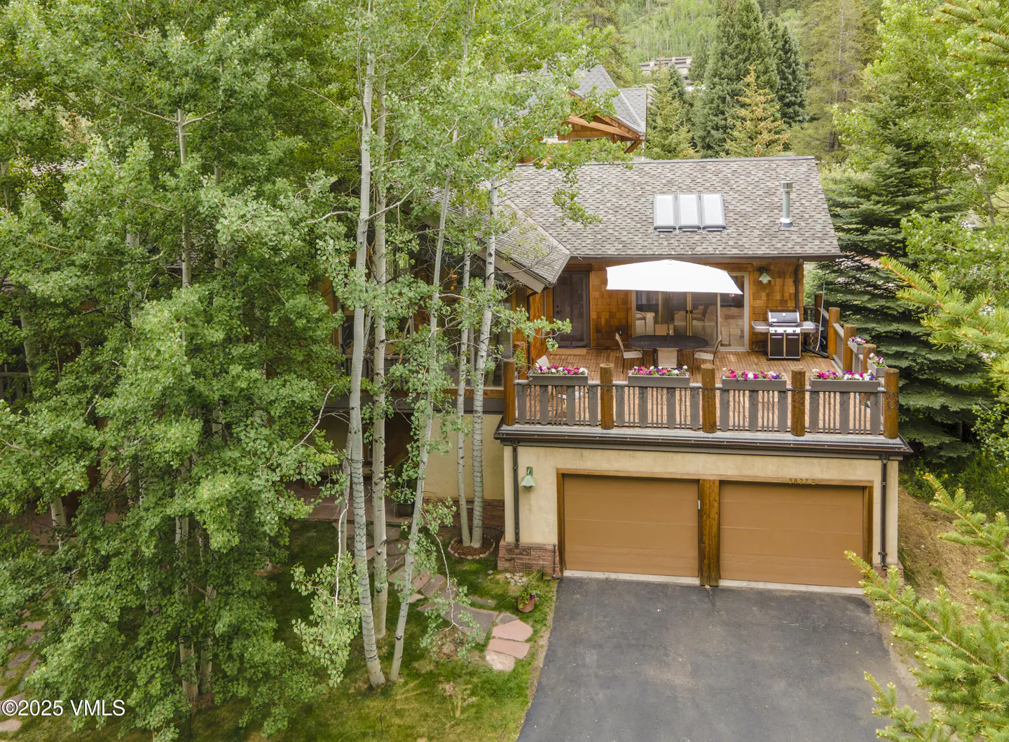 Two-story house with a brown garage, a deck with a white umbrella, and green trees.