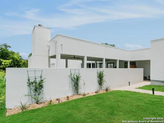 Modern white house with a flat roof, a covered patio, and a white wall with climbing plants in front of a green lawn.