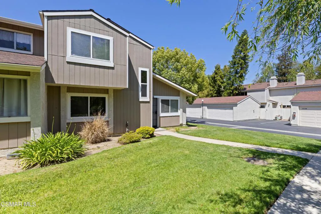 Exterior of a two-story, tan townhouse with green lawn, bushes, and white-framed windows under a clear blue sky. Garages are visible in the background.