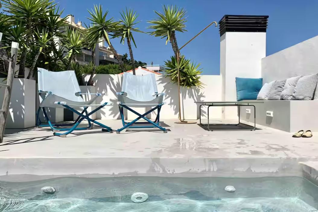 Sunny rooftop terrace with a small pool, two folding chairs, a bench with cushions, and palm trees against a blue sky.