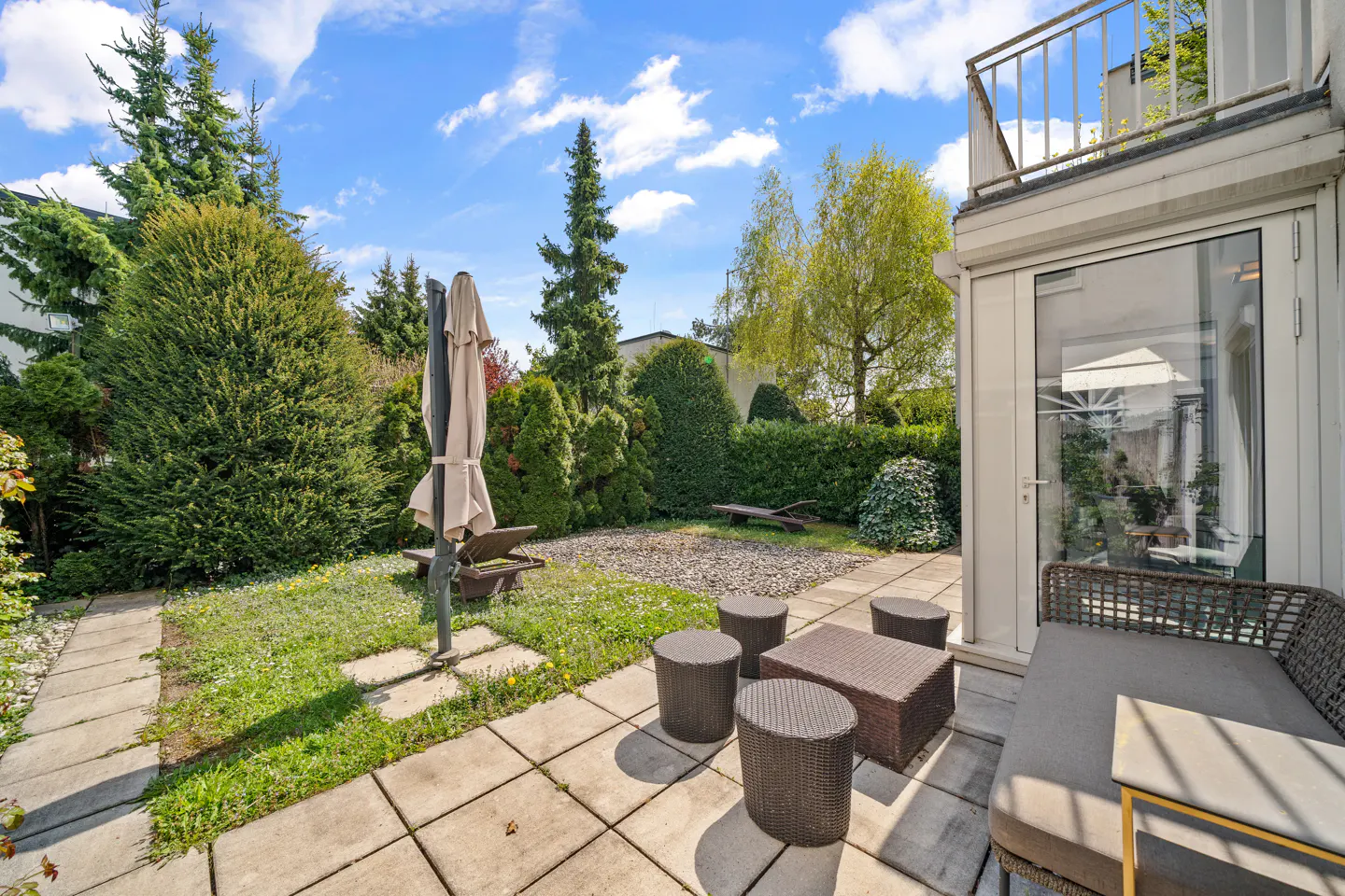 A backyard patio with a wicker table and chairs, a lounge chair, and a closed umbrella. Green trees and bushes surround the patio.