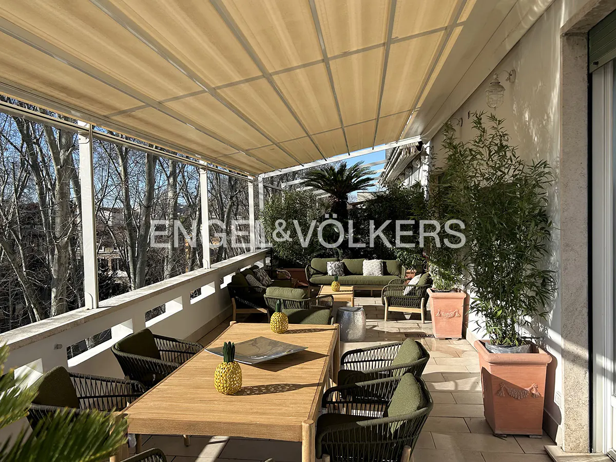 Spacious balcony with a wooden dining table, green wicker chairs, potted plants, and a large beige retractable awning.