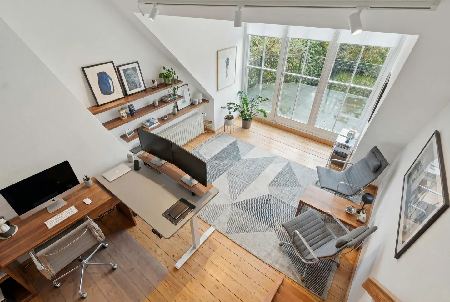Bright, modern home office with wooden floors, white walls, desks, chairs, and a large window overlooking greenery. Shelves display art and decor.