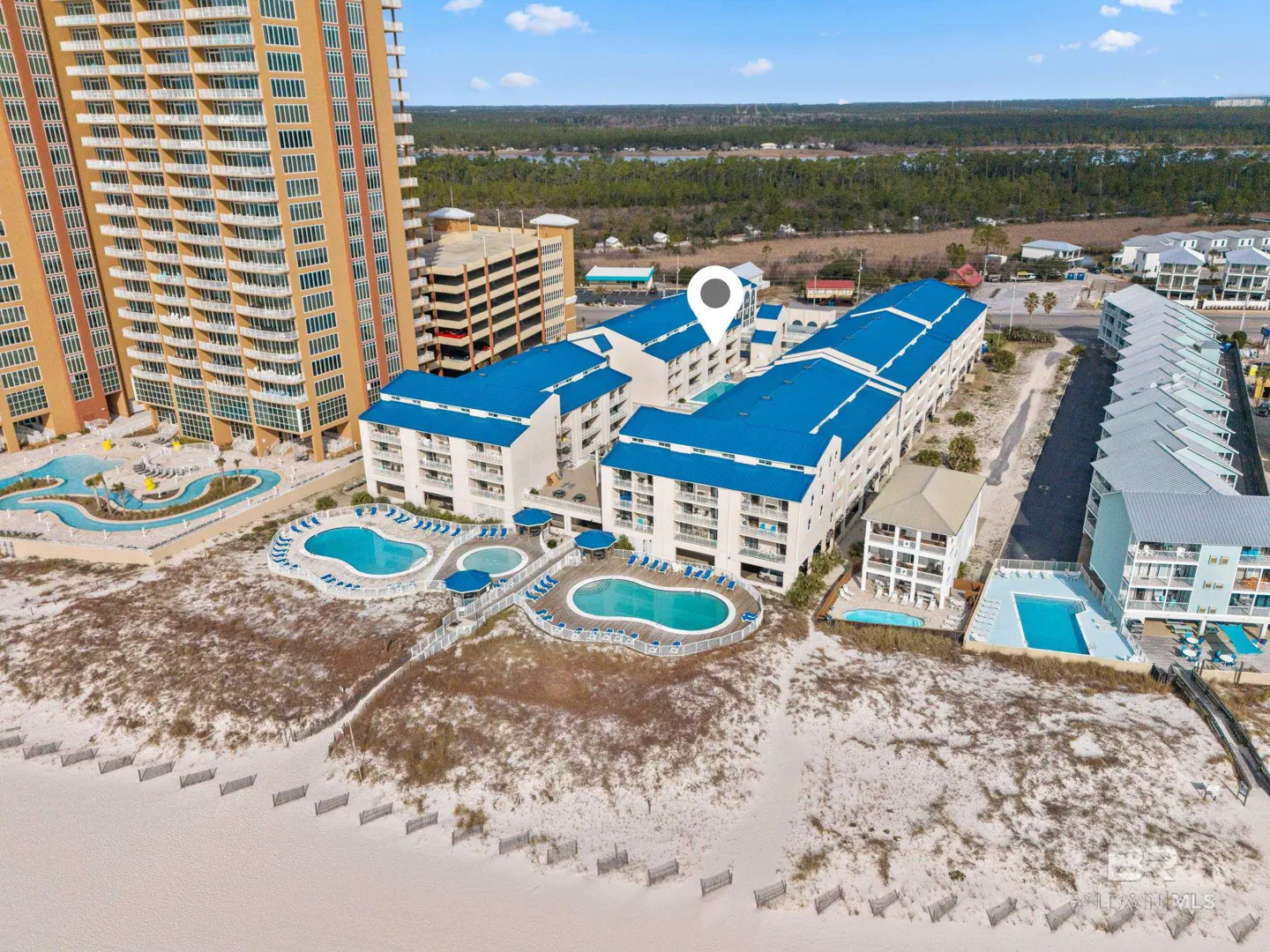 Aerial view of a beachfront condo complex with blue roofs, pools, and a tall orange building nearby.