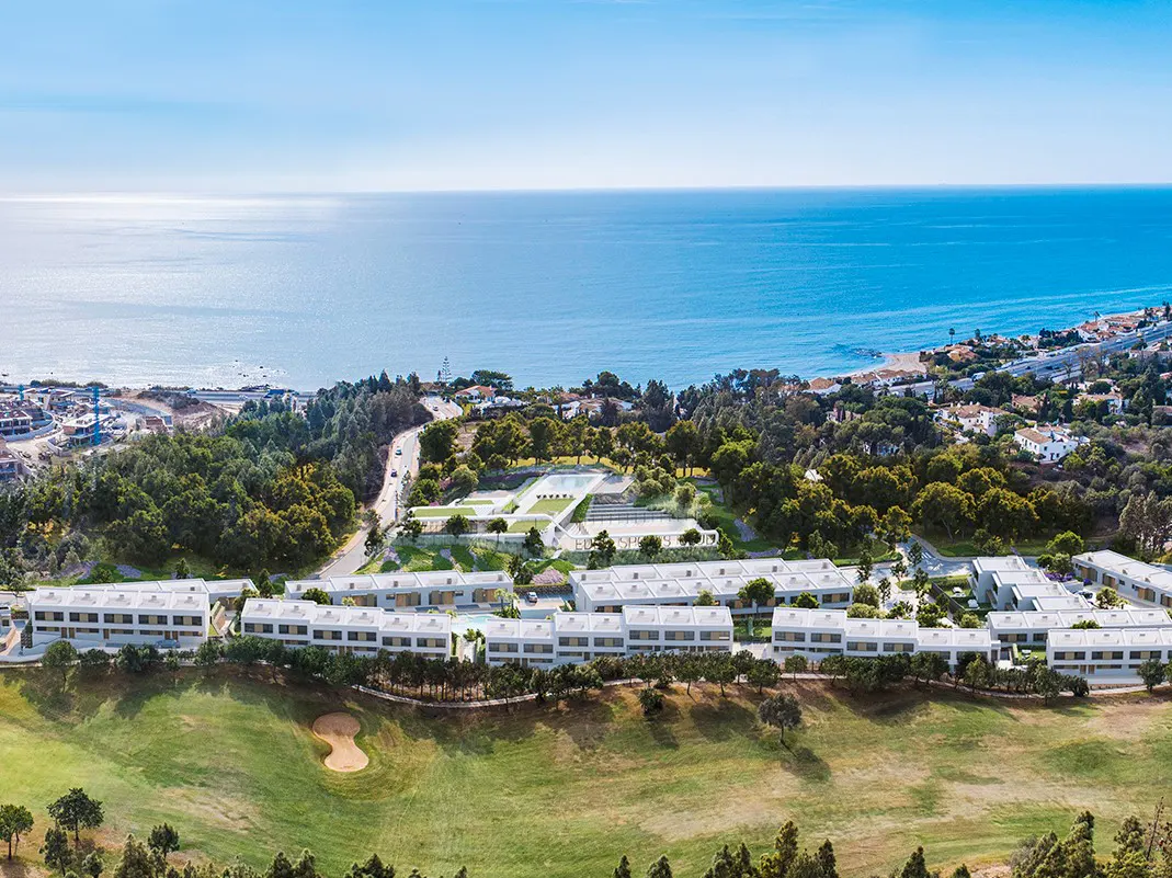 Aerial view of modern white townhouses on a green golf course near the ocean under a blue sky.