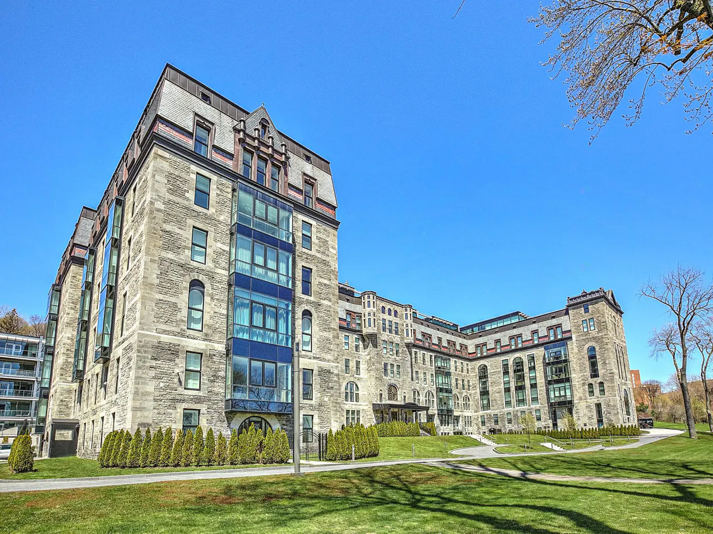 Exterior view of a large, grey stone apartment building with blue glass balconies and a green lawn under a clear blue sky.
