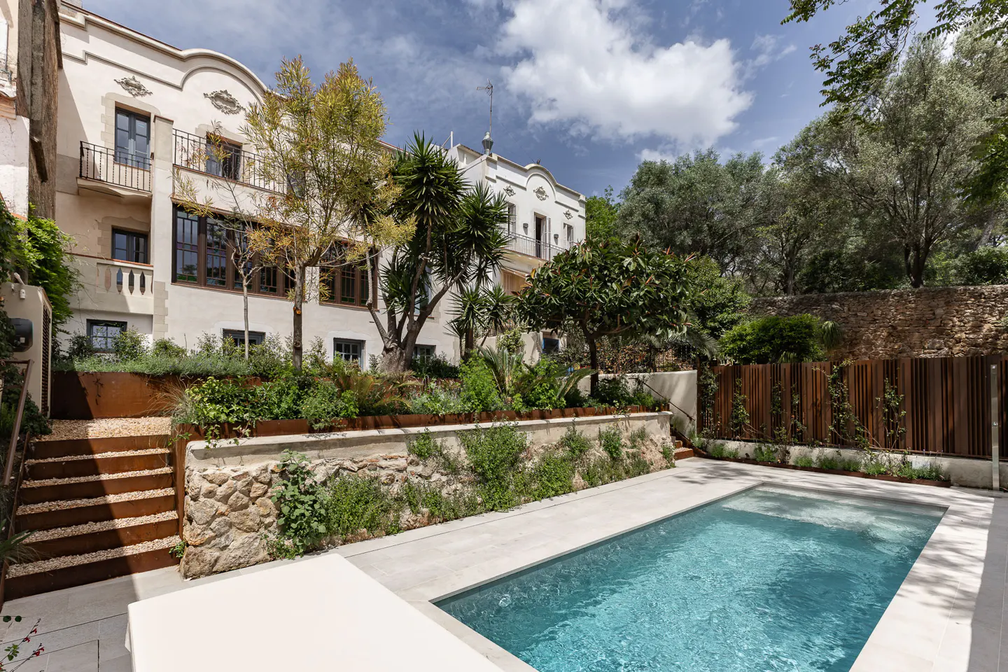 A backyard with a pool and a two-story white building with brown window frames and balconies. The pool is rectangular and has light blue water.
