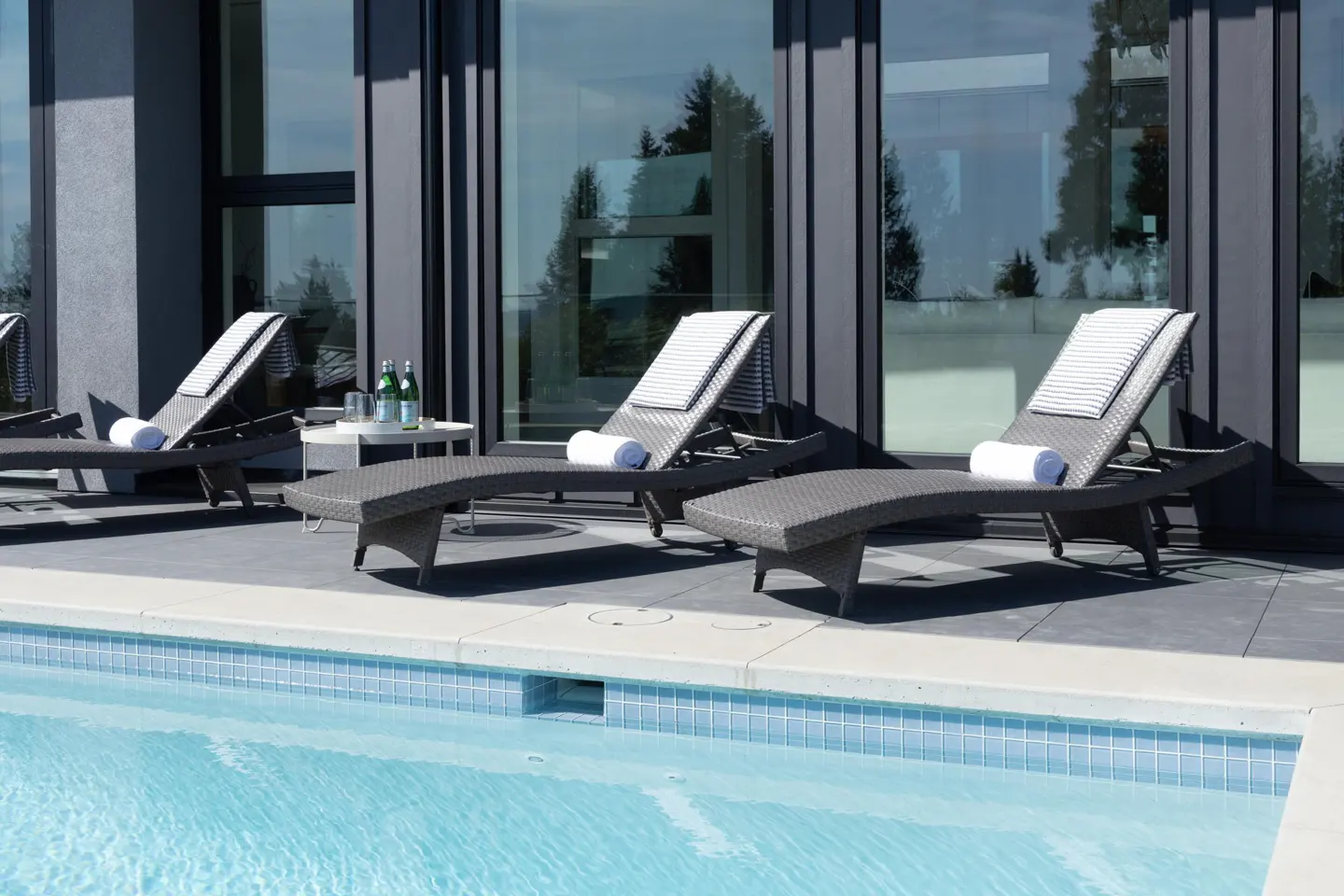 Outdoor pool area with three lounge chairs, white towels, and a small table with drinks. Modern building with large windows in the background.