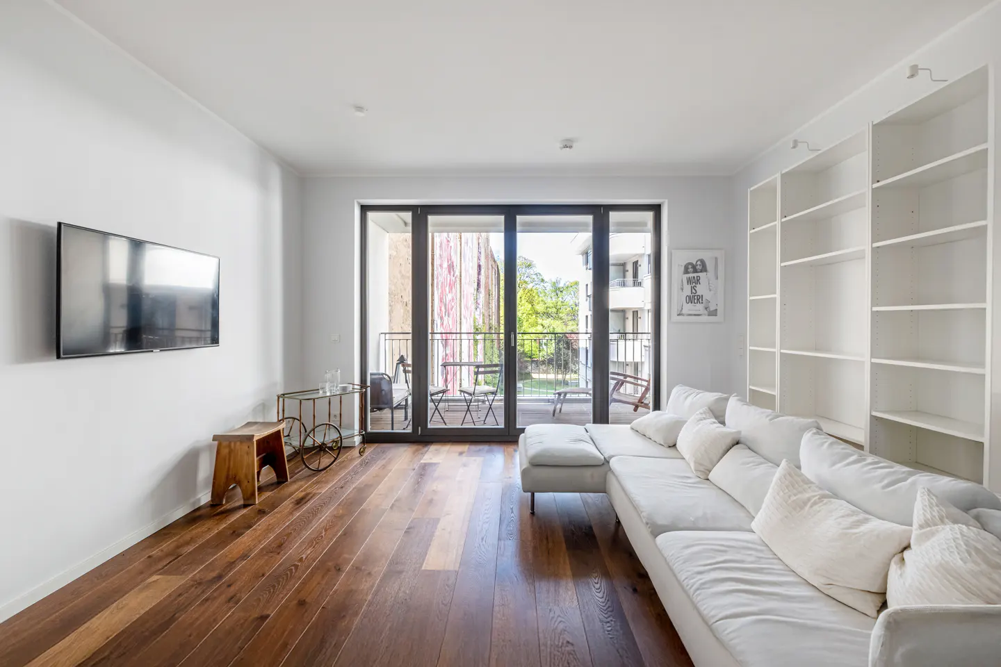 Bright living room with hardwood floors, white walls, and a large white sofa. A TV hangs on the wall, and a balcony is visible through glass doors.