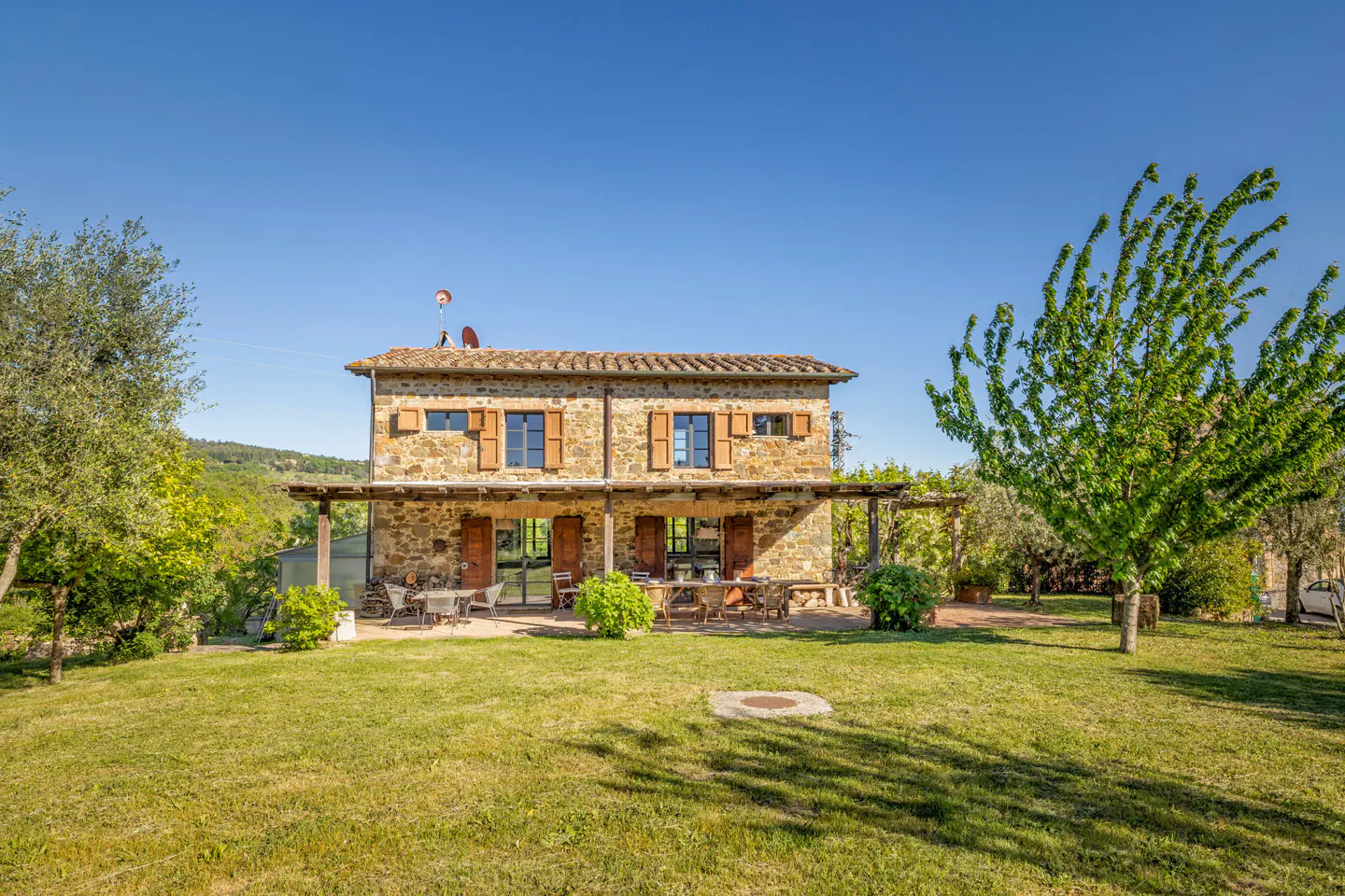 Two-story stone house with brown shutters and a tile roof, set on a green lawn under a clear blue sky.