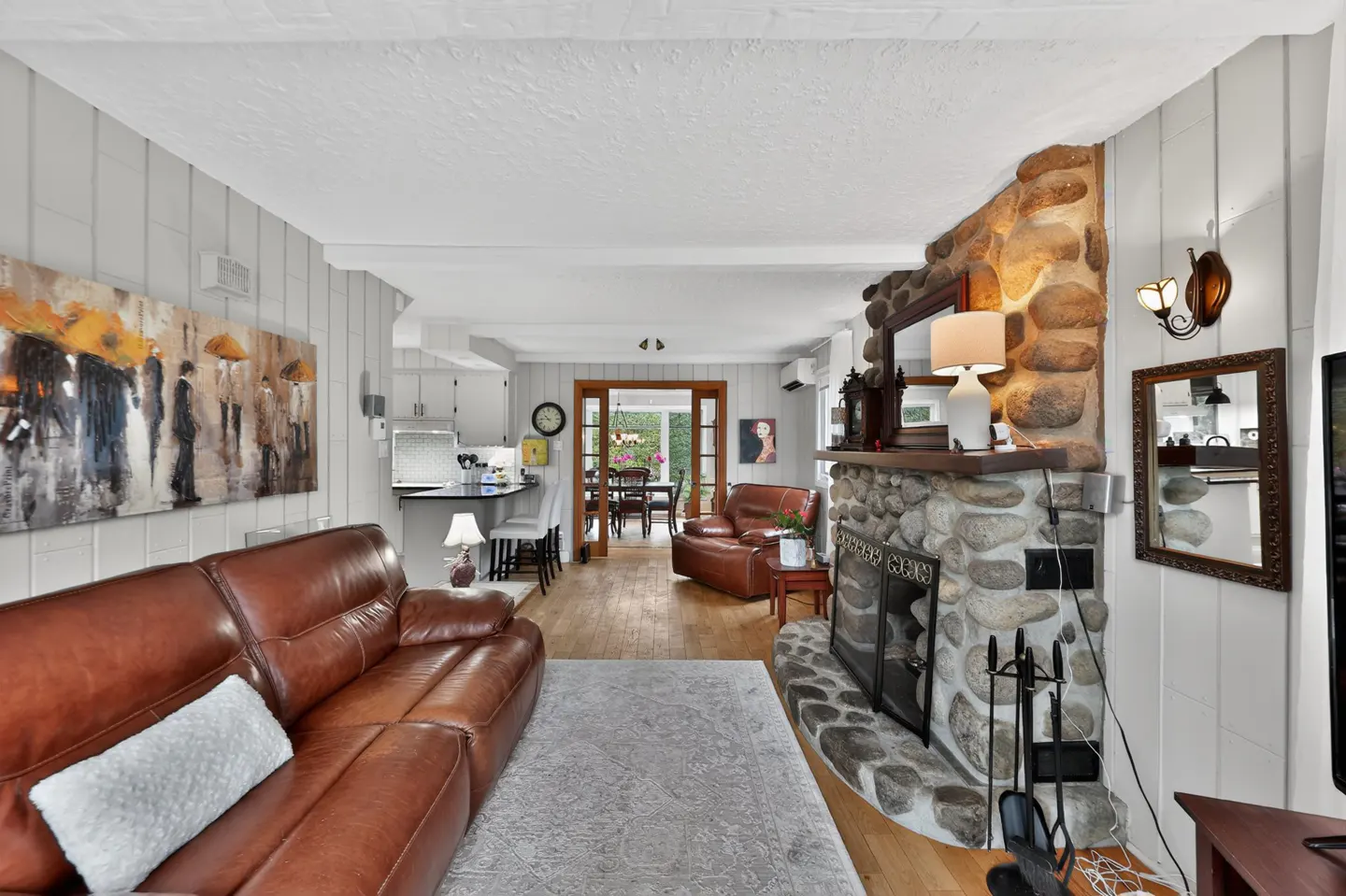 Living room with a brown leather sofa, stone fireplace, and open floor plan to the kitchen and dining area. A painting hangs on the wall.