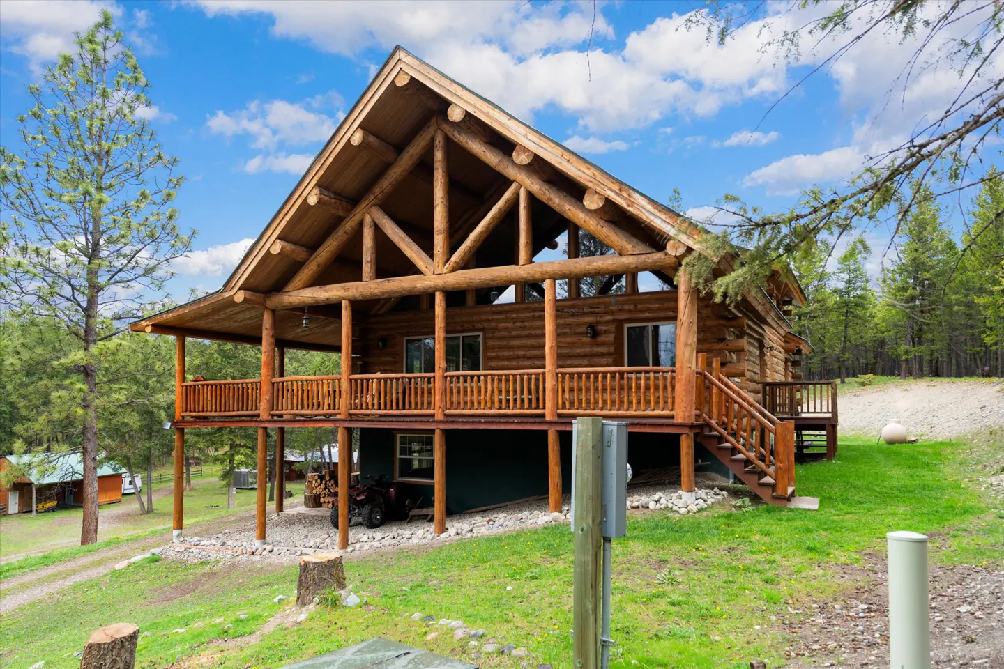 Exterior view of a two-story log cabin with a wooden porch and stairs, set on a grassy hill under a blue sky with clouds.