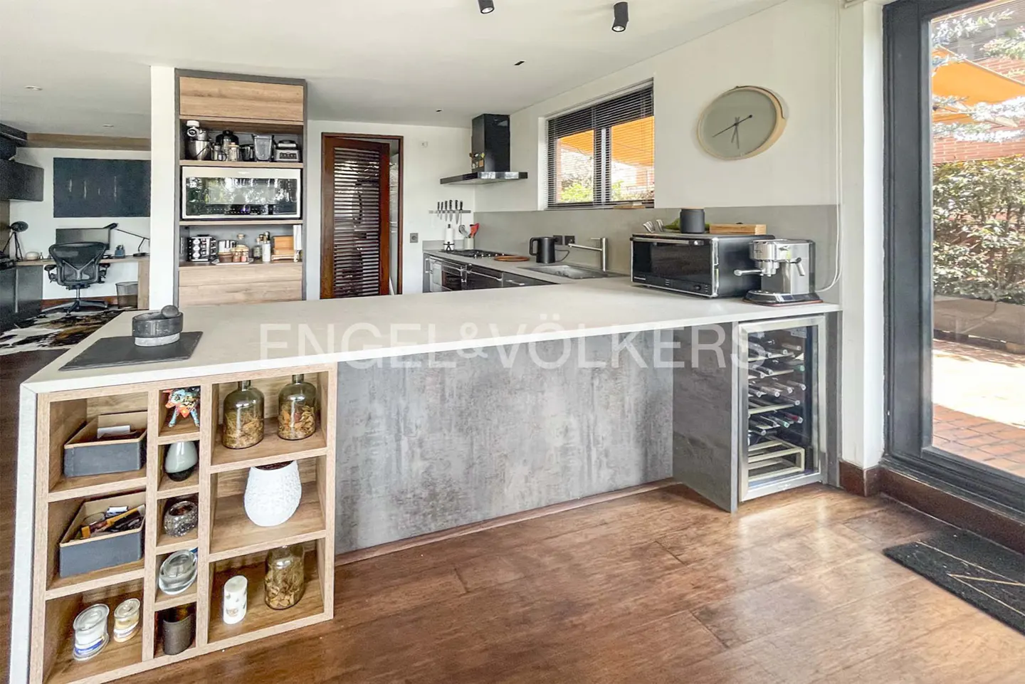 A modern kitchen with a large island, wooden floors, and a wine cooler. Natural light streams in from a nearby window.