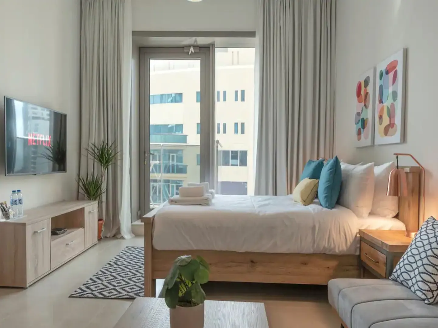 Modern bedroom with a wooden bed, white linens, colorful pillows, a TV, and a balcony view of city buildings.