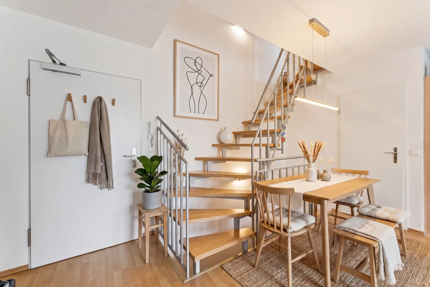 Bright apartment interior with wooden stairs, dining table, and white walls. A tote bag and scarf hang near the entrance door.