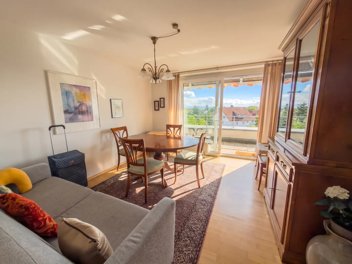 Bright living room with a gray sofa, round wooden table with chairs, and a balcony with a view of houses and trees.